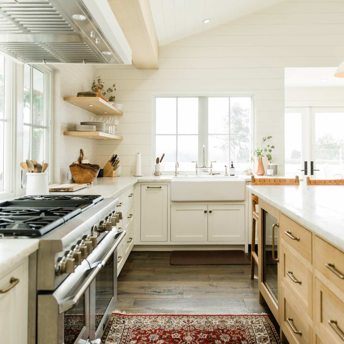 A modern kitchen with a large island, white cabinetry, wooden shelving, and large windows illuminating the space.