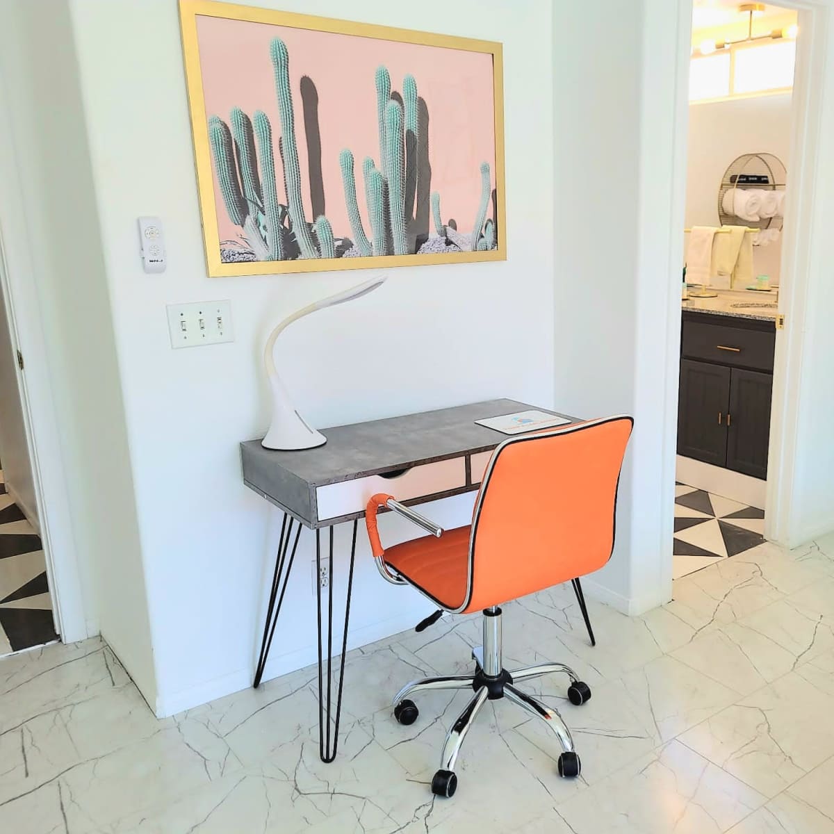 A modern workspace featuring a gray desk with an orange chair, a cactus-themed wall art, and a view into a bathroom through an open doorway.