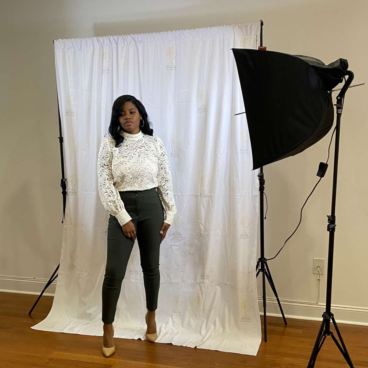 A woman in a white lace top and green pants stands in front of a white backdrop with studio lighting setup.