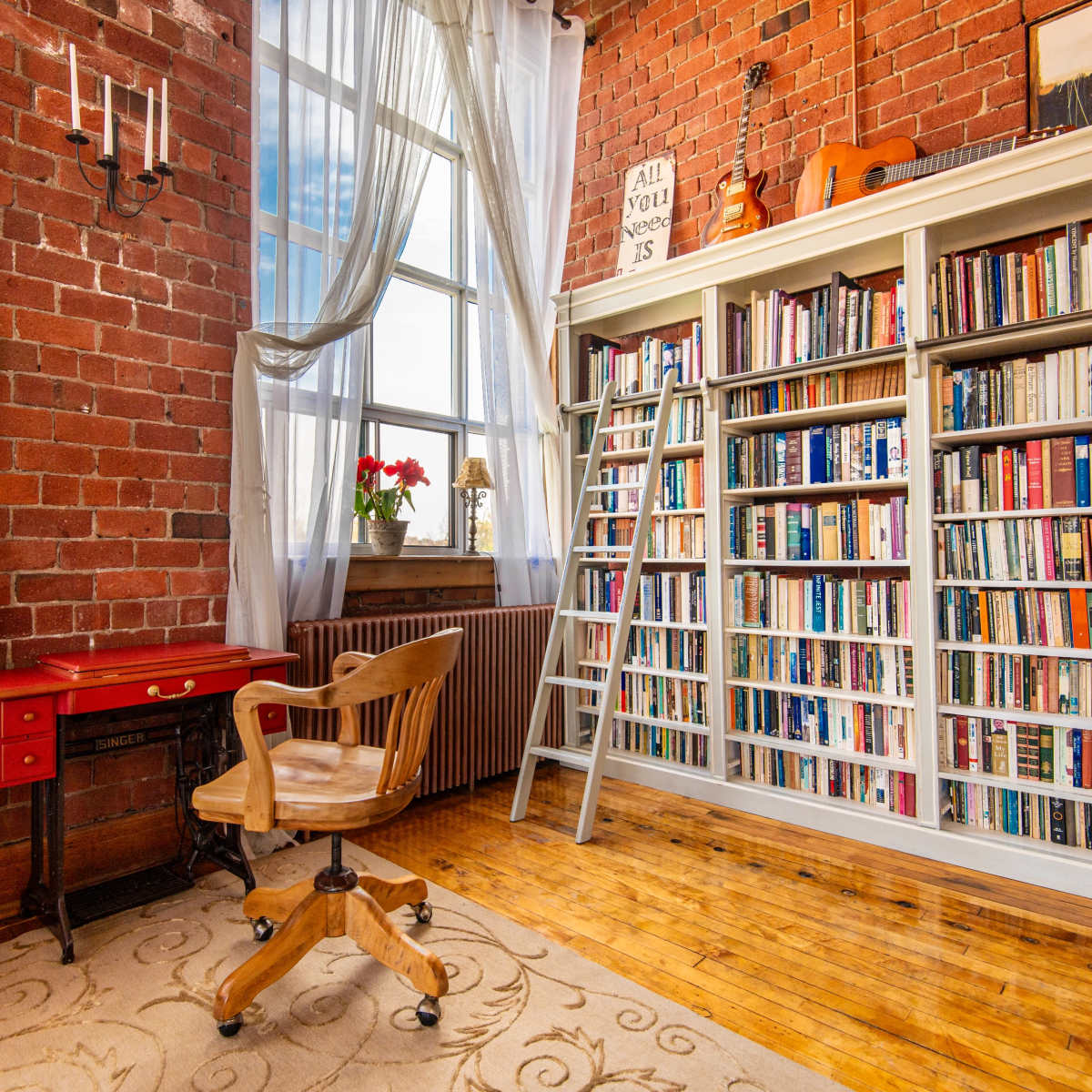 A cozy reading nook features a wooden bookcase filled with books, a red desk with a chair, and large windows with sheer curtains.