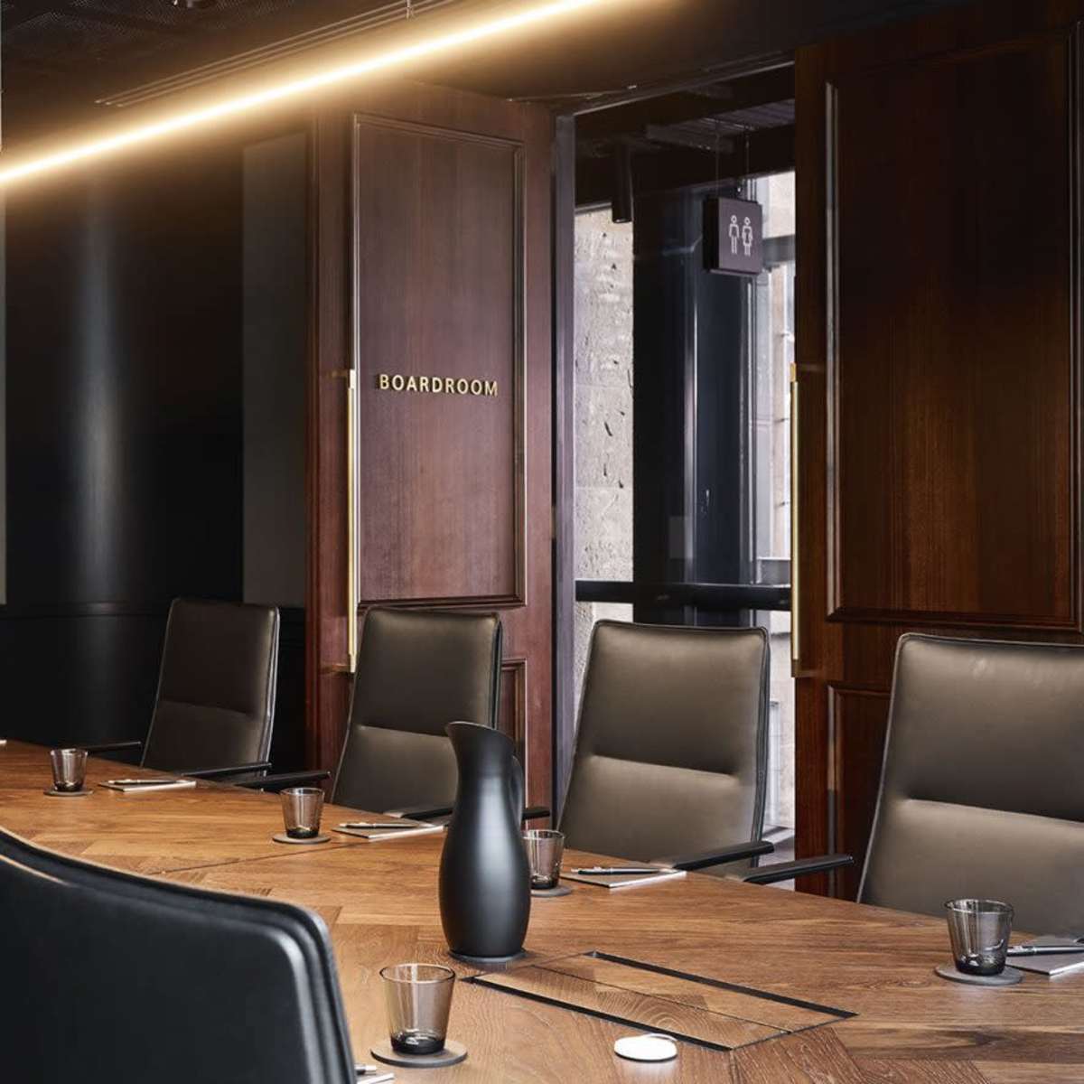 A modern boardroom with a long wooden table, black chairs, a large screen on the wall, and a door labeled "BOARDROOM."