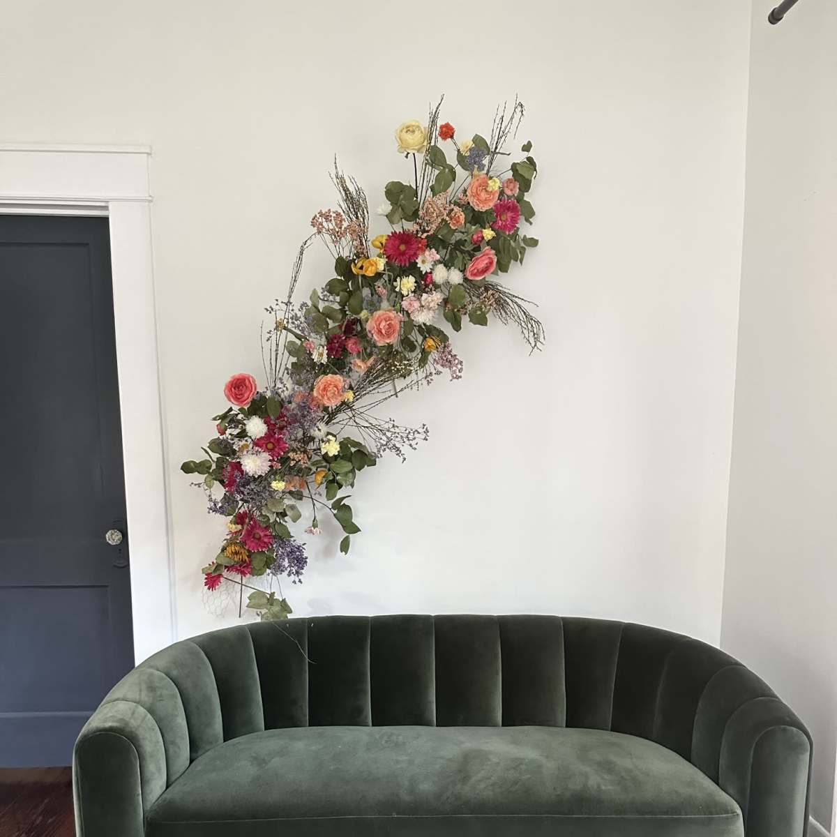 A green velvet sofa is positioned against a white wall adorned with an arrangement of colorful flowers and greenery.