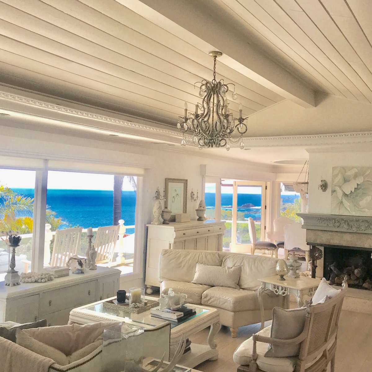 A bright living room features white furniture, a chandelier, and large windows overlooking the ocean.