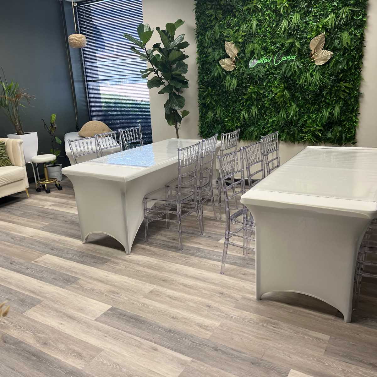 A modern room with two white tables, clear chairs, a sofa, and greenery on the walls.
