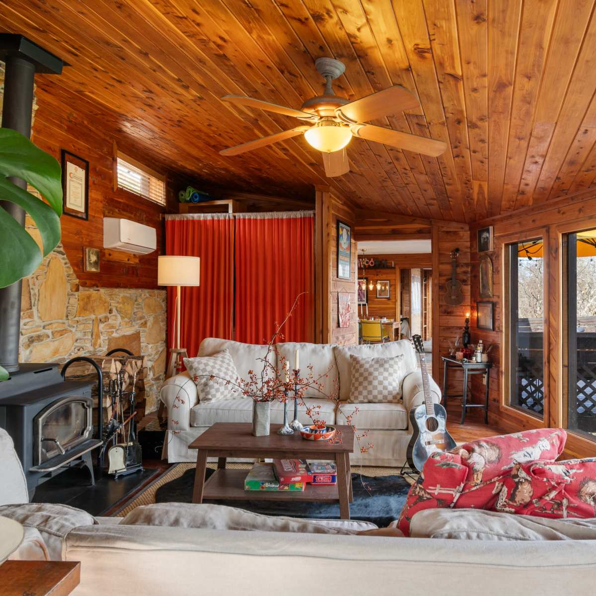 A cozy living room with wooden walls and ceiling, featuring a sofa, armchairs, a coffee table, a fireplace, and large windows leading to an outdoor area.