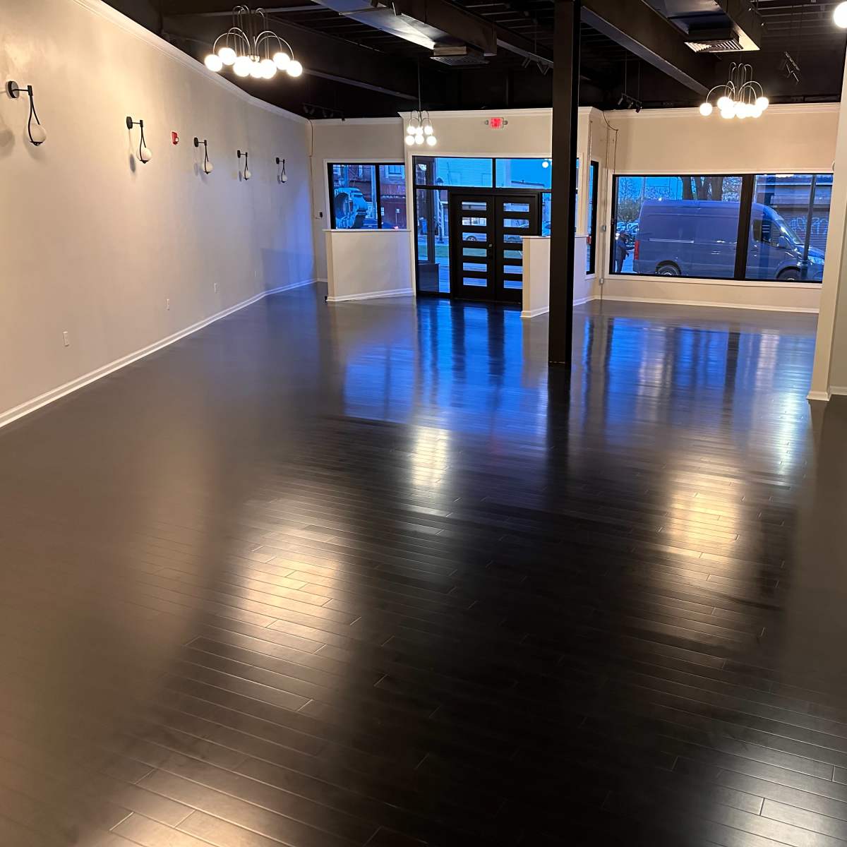 A spacious, empty room with dark wooden floors, large windows, and multiple light fixtures hanging from the ceiling.