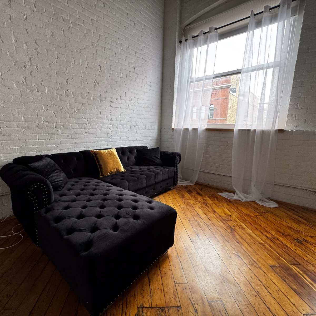 A black tufted sectional sofa with a yellow cushion is positioned in a room with white brick walls and a large window draped in sheer curtains.
