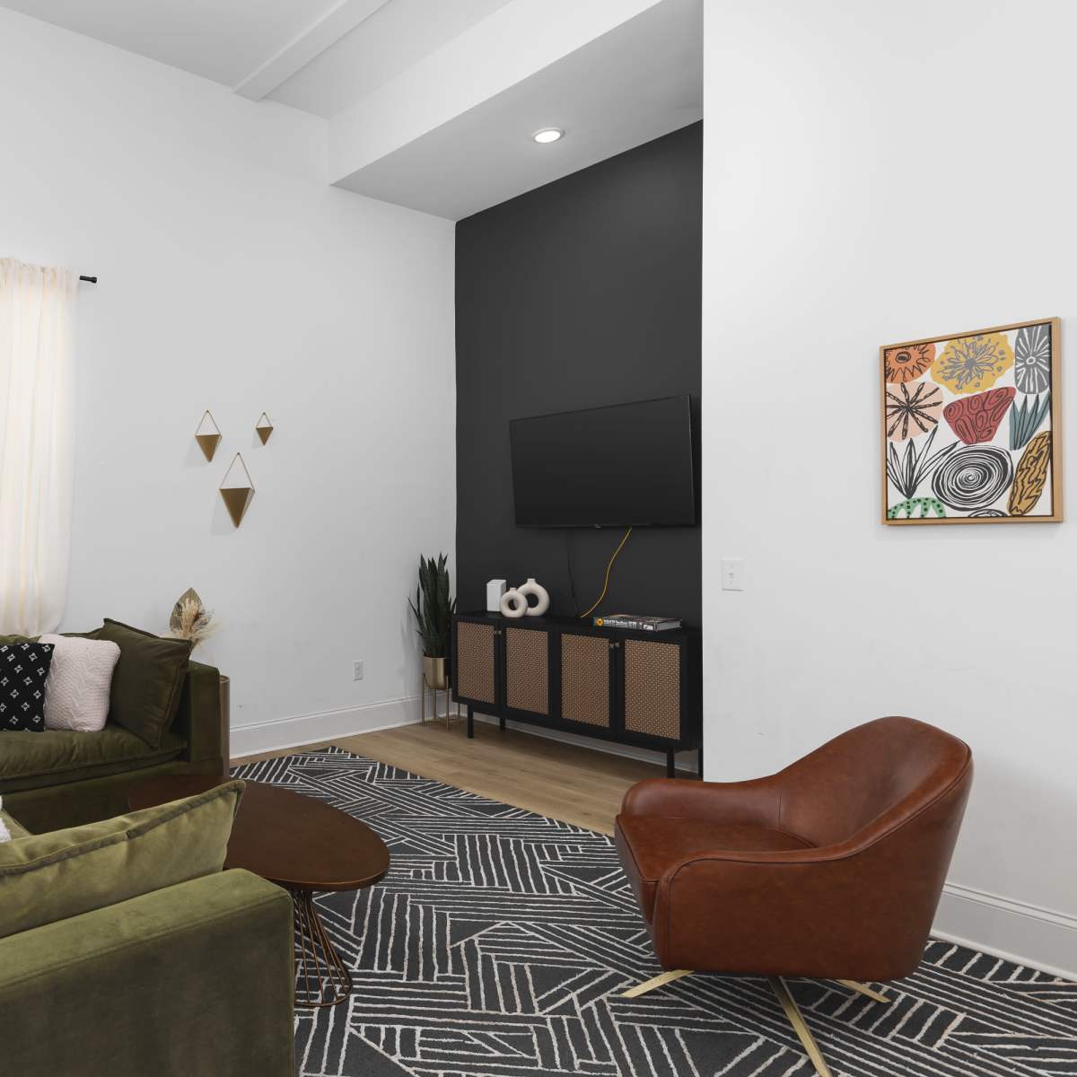 A modern living room features a green sectional sofa, a round coffee table, and a brown accent chair, with a TV mounted on a dark accent wall and decorative art pieces around the space.