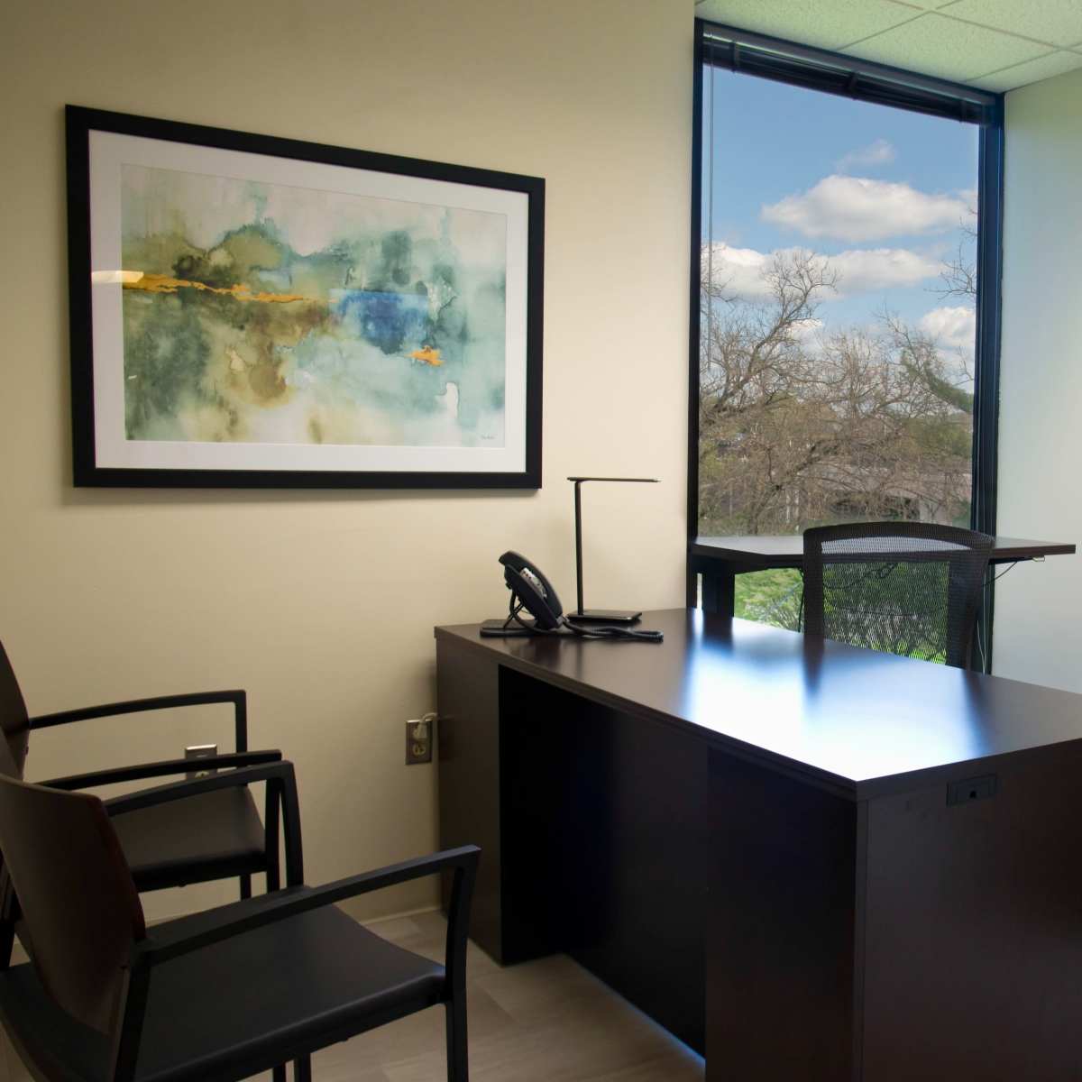 A modern office features a wooden desk, two chairs, a phone, and a large window showcasing a view of trees and a blue sky.