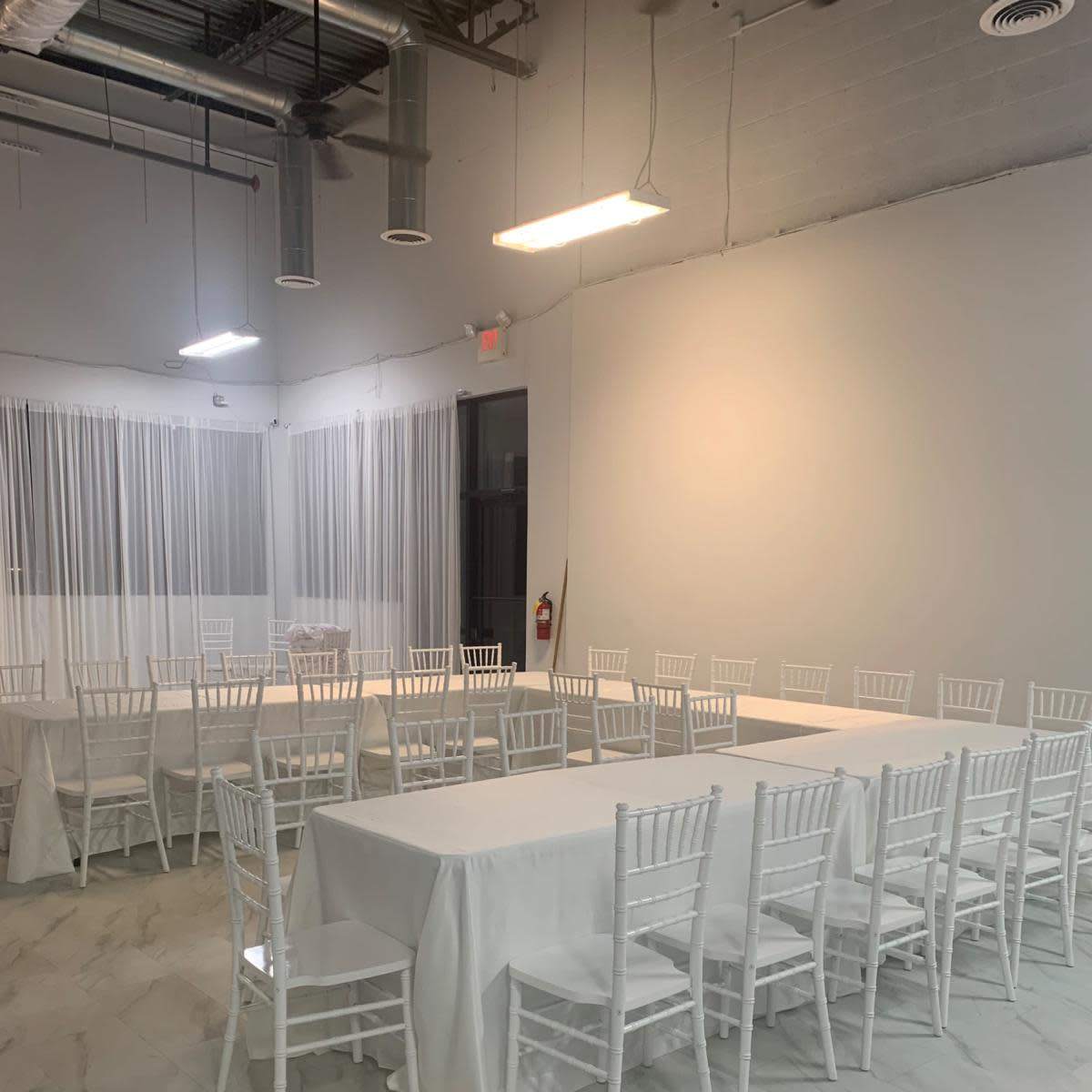 A spacious room with two long tables covered in white tablecloths, surrounded by white chairs, and illuminated by overhead lights.