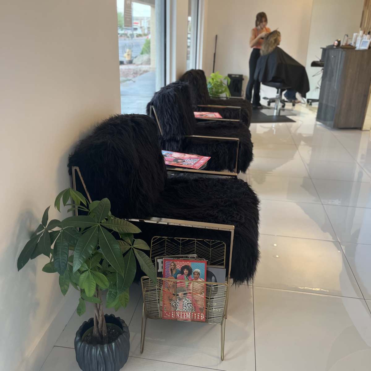 A modern hair salon with black chairs, a plant, and a stylist working on a client in the background.