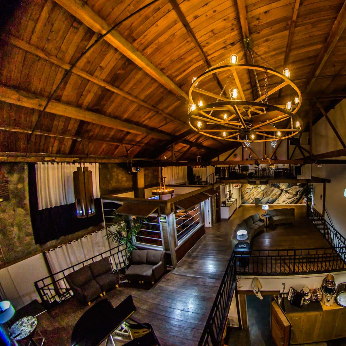 The interior of a spacious loft features exposed wooden beams, a large circular chandelier, and various seating areas arranged around a central open space.