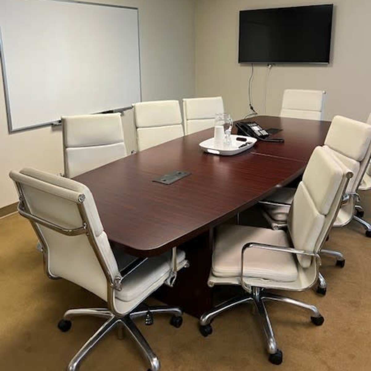 A conference room with a large wooden table surrounded by eight white chairs, a flat-screen TV mounted on the wall, and a whiteboard.