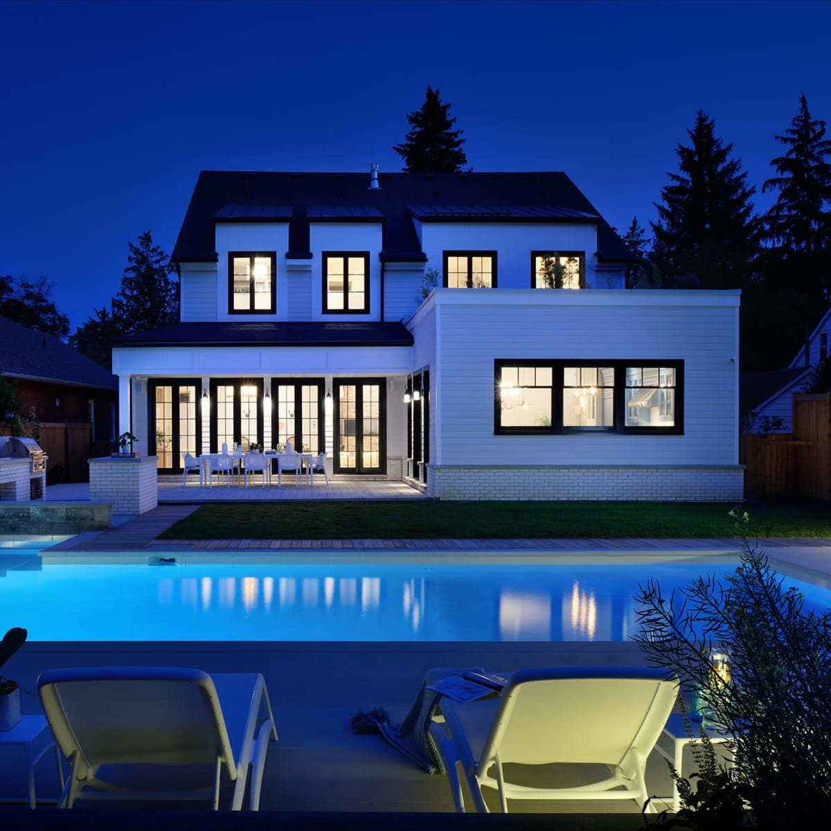 A modern house with large windows overlooks a swimming pool illuminated in soft blue light during the night.