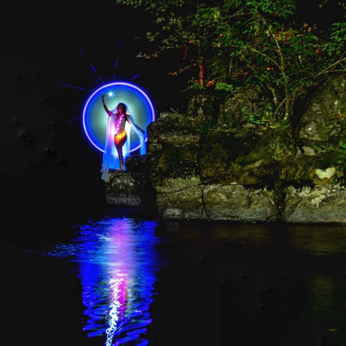 A figure stands on a rock by a calm body of water, illuminated by colorful light against a dark backdrop.