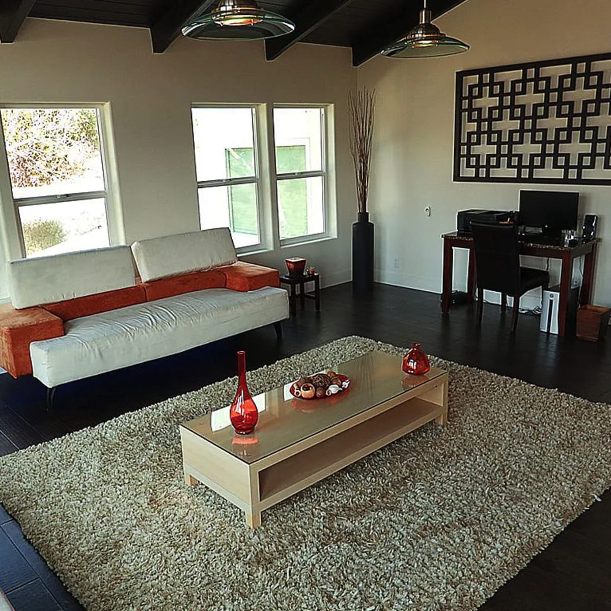 A furnished living room featuring a white and orange sofa, a coffee table on a large area rug, and a desk with a computer near large windows.