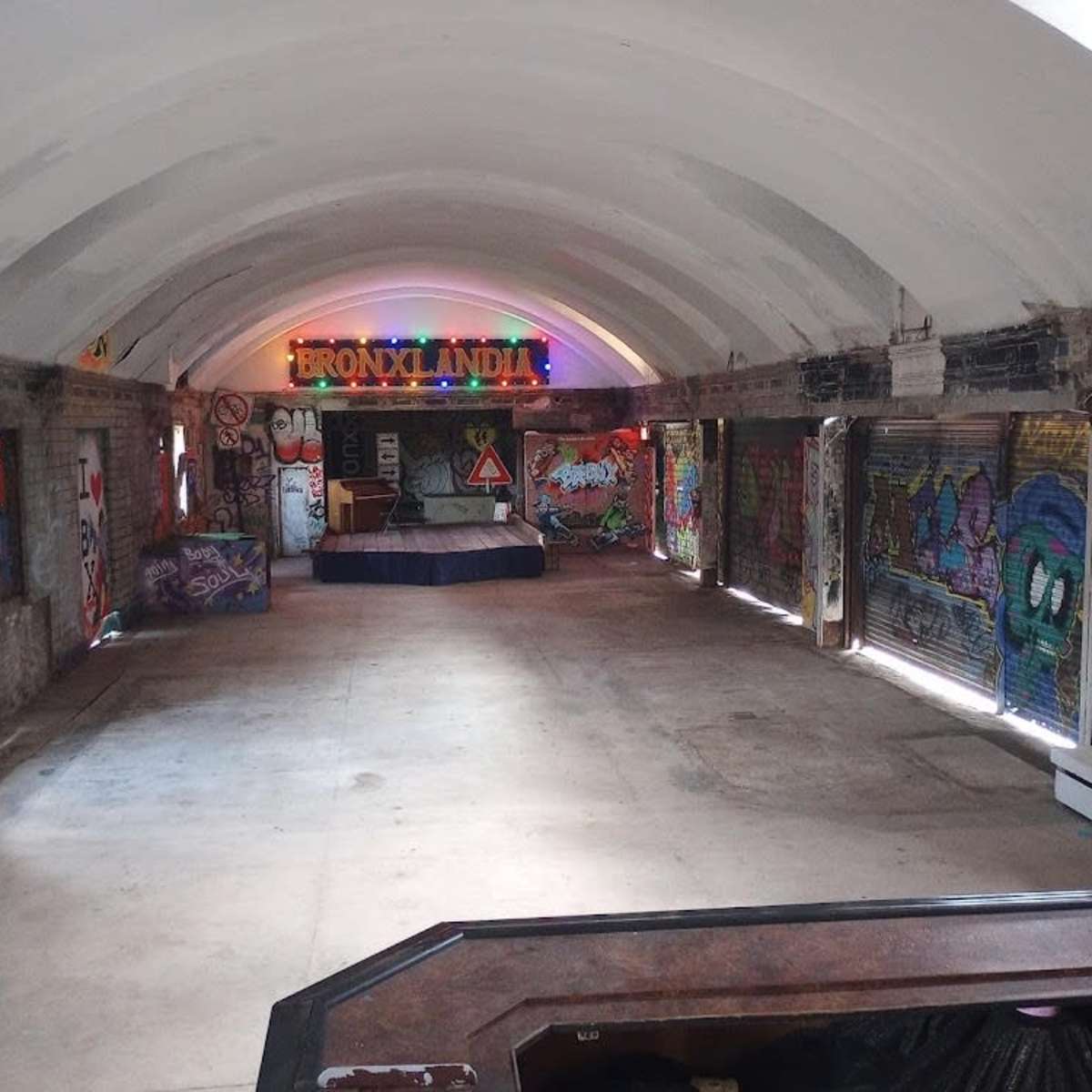 An abandoned indoor space with colorful graffiti on the walls and a stage at one end, illuminated by neon lights reading "Bronxlandia."