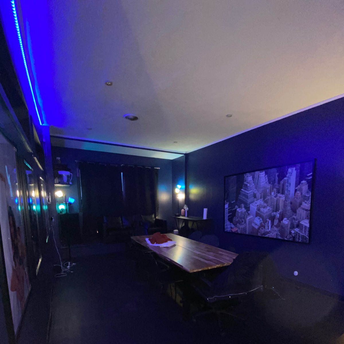 A dimly lit room with a large table, dark seating, and a wall-mounted screen displaying a cityscape.