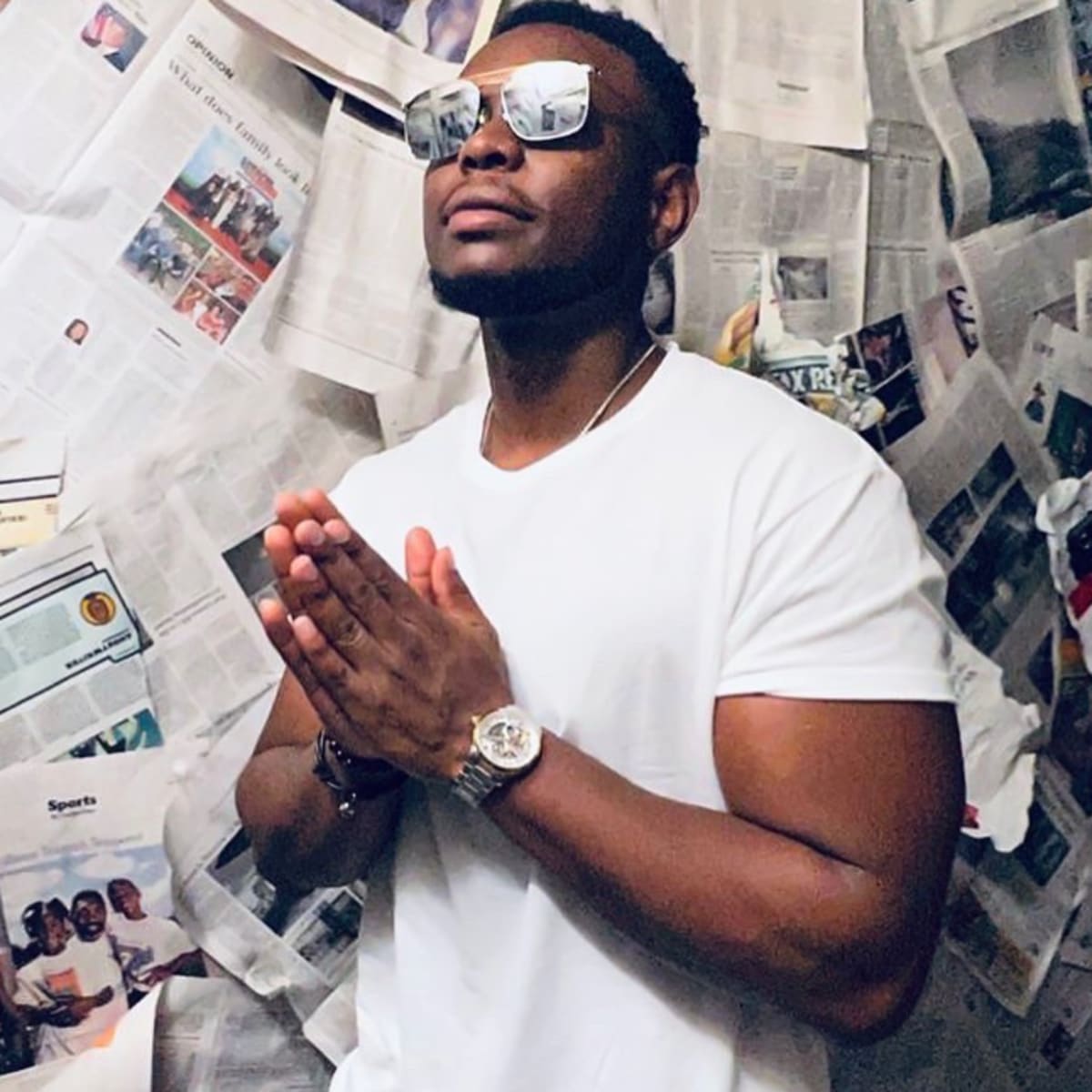 A man in a white t-shirt and sunglasses stands with his hands pressed together in a room covered in newspapers.