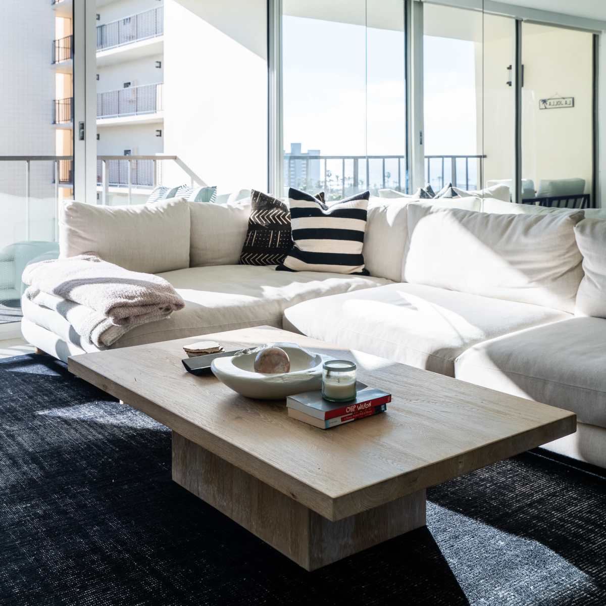 A spacious, modern living room features a large sectional sofa, a wooden coffee table with décor items, and expansive windows letting in natural light.