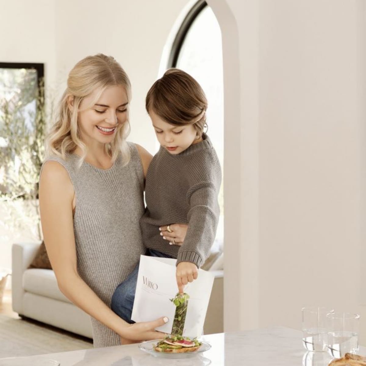 A woman holds a container while a young boy reaches inside to grab some greens on a marble kitchen counter with food plates arranged nearby.