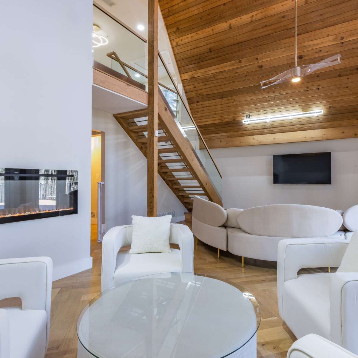 A modern living room features a circular sofa, glass coffee table, and a linear fireplace, with a staircase leading to an upper level.