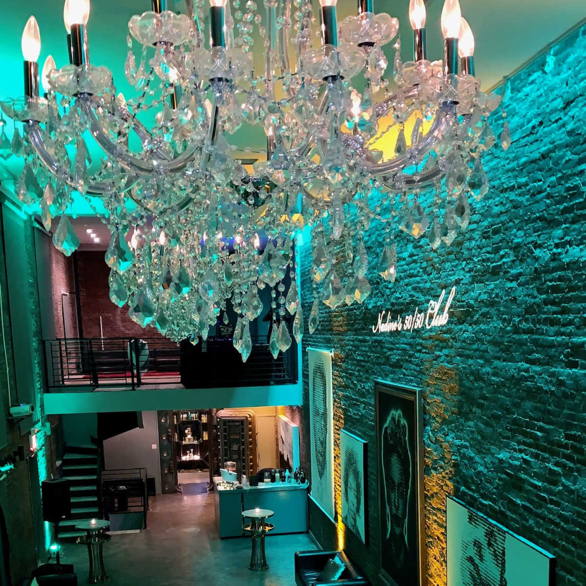 A spacious interior with a large chandelier, brick walls, seating areas, and artistic portraits illuminated by colored lighting.