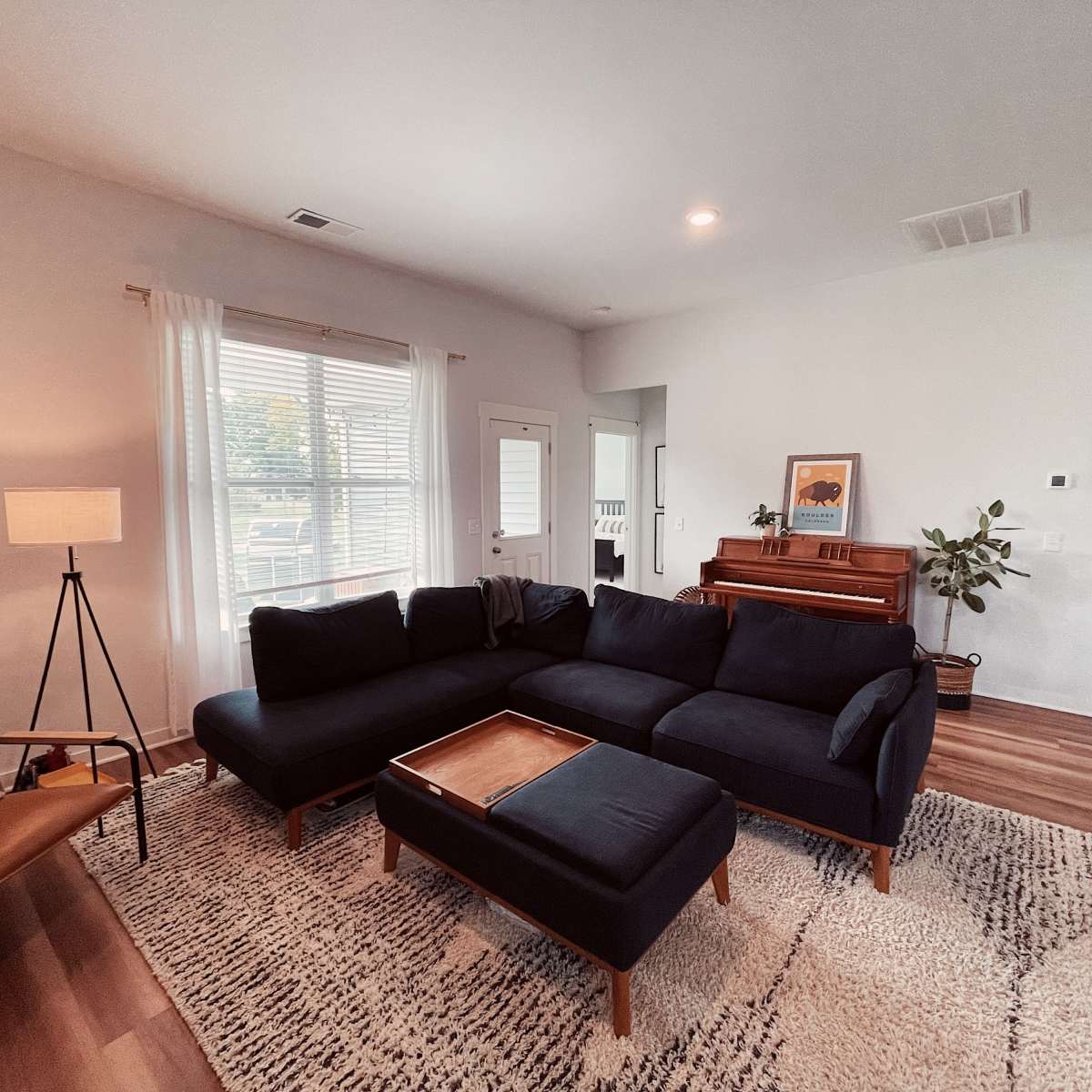 A living room features a dark sectional sofa, a wooden coffee table, a lamp, and a piano, with windows allowing natural light to enter.