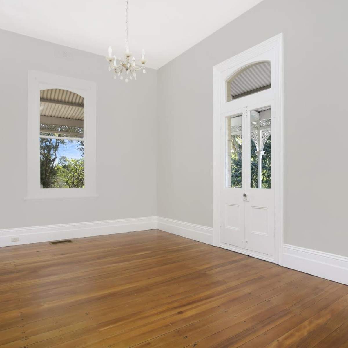 The room features light gray walls, wooden flooring, a chandelier, and large windows with a door leading outside.