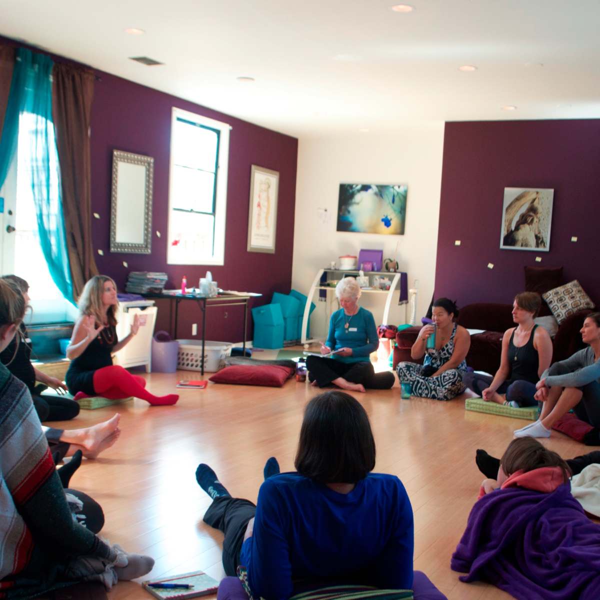 A group of people sit in a circle on the floor of a room with purple walls, engaged in a discussion.