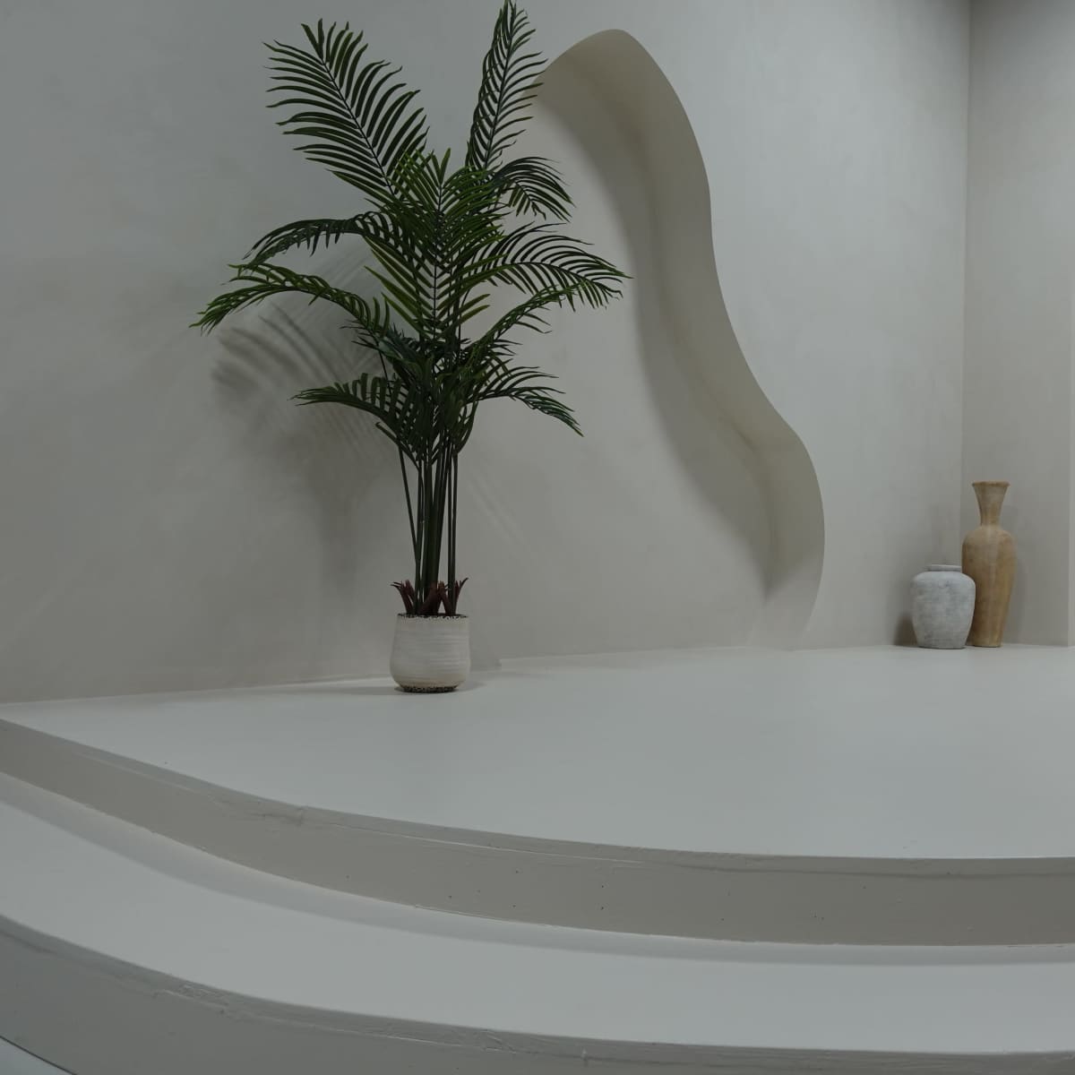 A minimalist interior space with smooth white walls, curved architectural features, and a potted palm tree beside decorative vases.