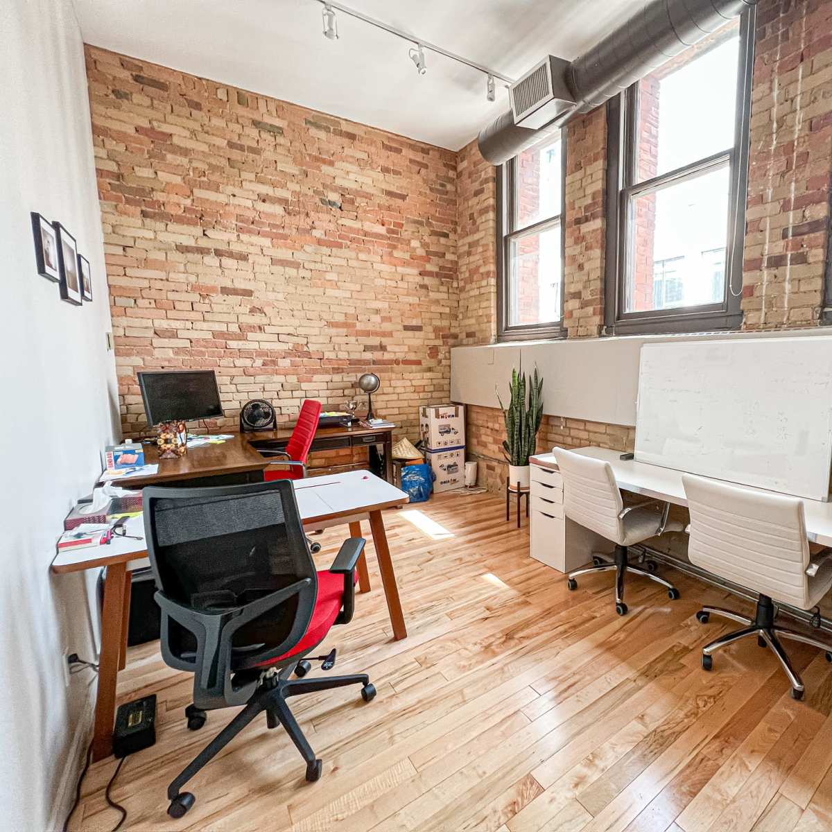 A modern office space with exposed brick walls, three desks, and large windows providing natural light.