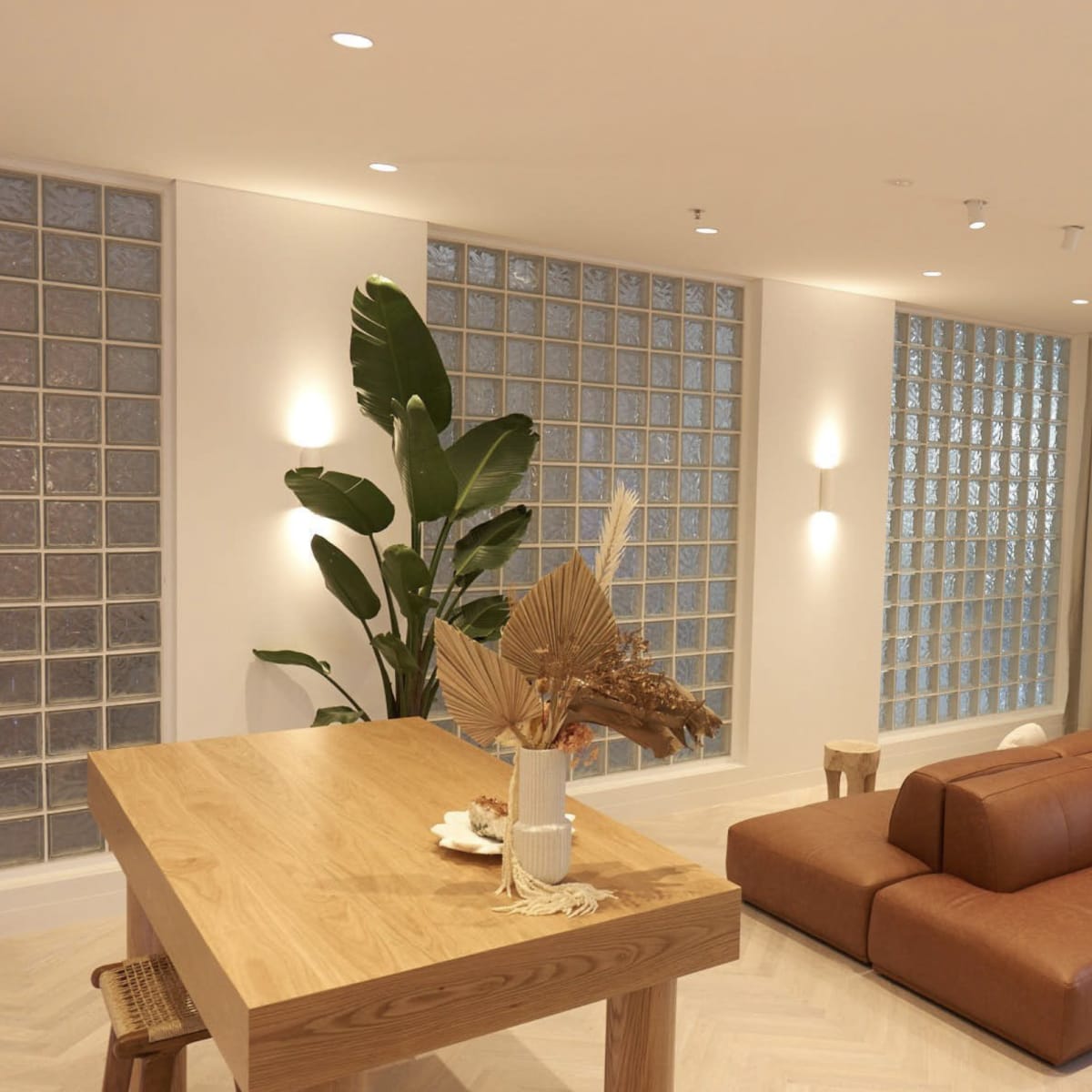 A modern interior space featuring a brown leather sofa, a wooden dining table, large glass block windows, and a circular mirror on the wall.