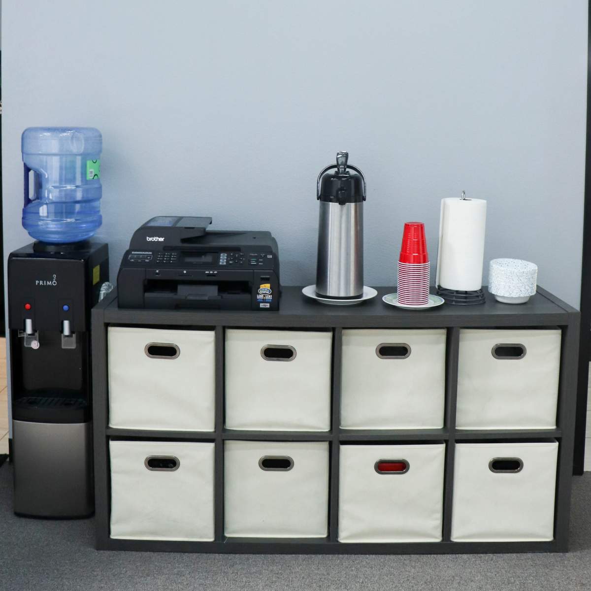 An office storage unit with fabric bins, a printer, a water cooler, a metal coffee pot, and a stack of red plastic cups.