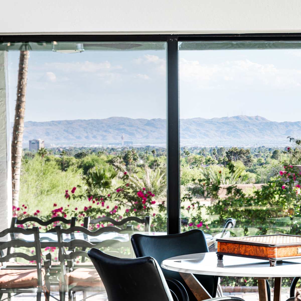 A modern dining area features a round table with a game board, overlooking flowering plants and distant mountains through large windows.
