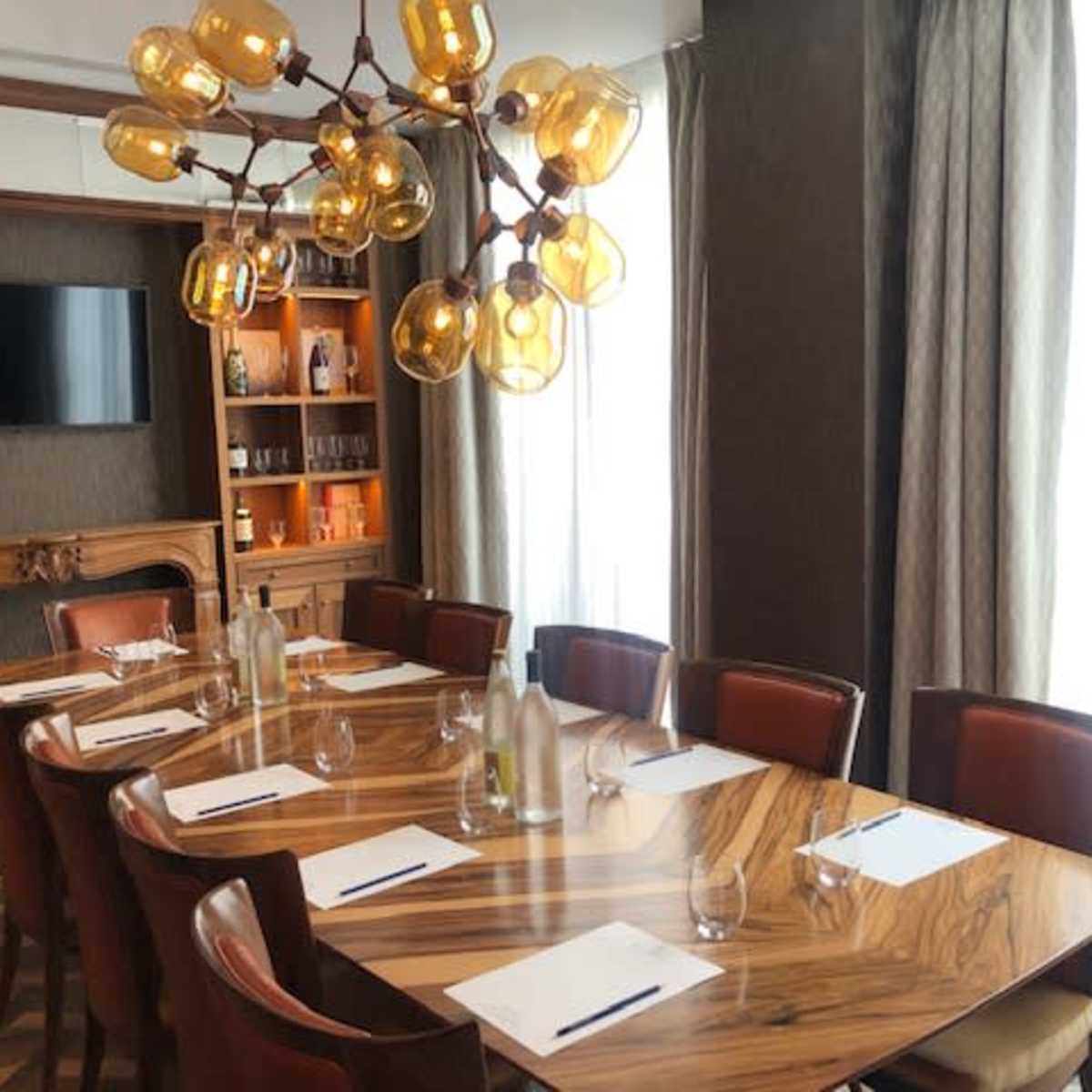 A well-lit conference room features a large wooden table set with glasses and stationery, surrounded by chairs and illuminated by a unique chandelier.