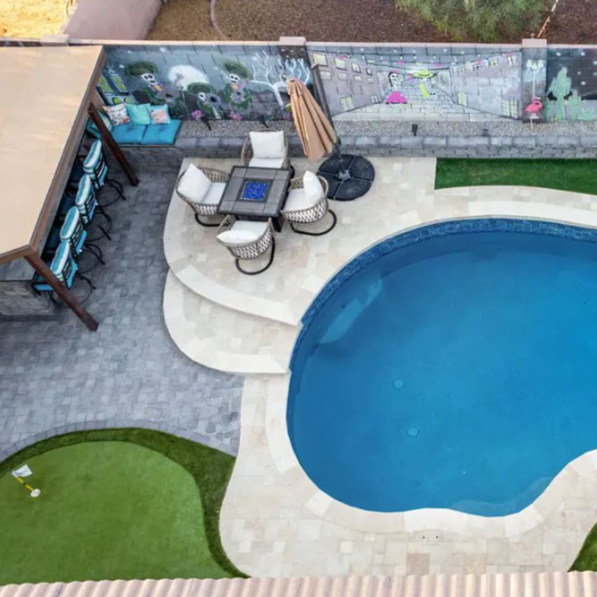 An aerial view of a swimming pool surrounded by a patio, lounge chairs, and an umbrella, with a mural-covered wall in the background.