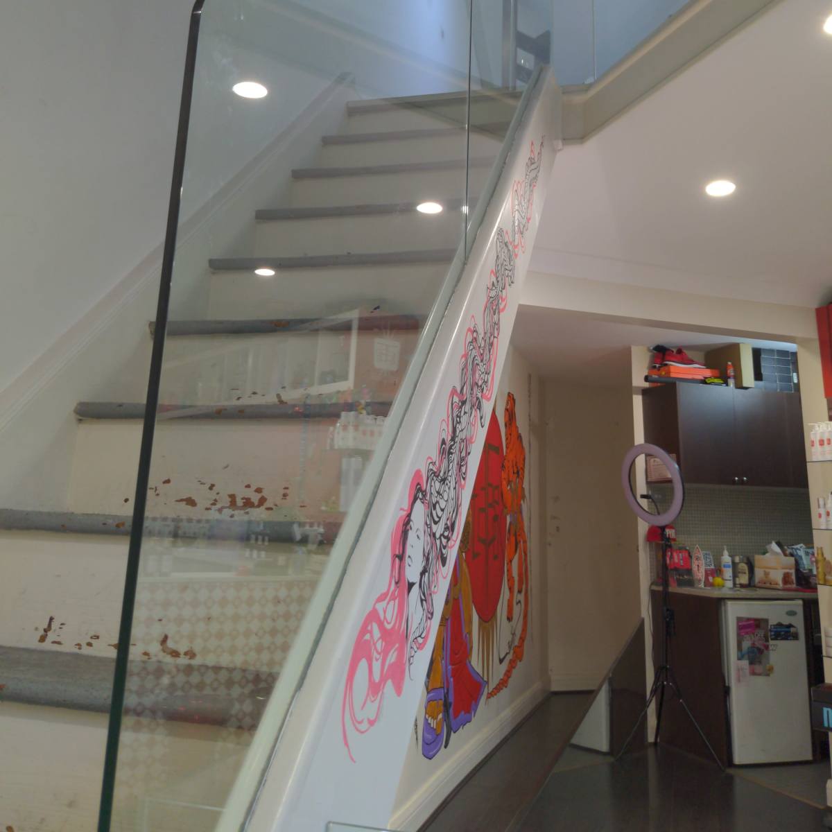 A glass-railed staircase leading to an upper level, with colorful artwork on the wall and a well-lit space at the top.