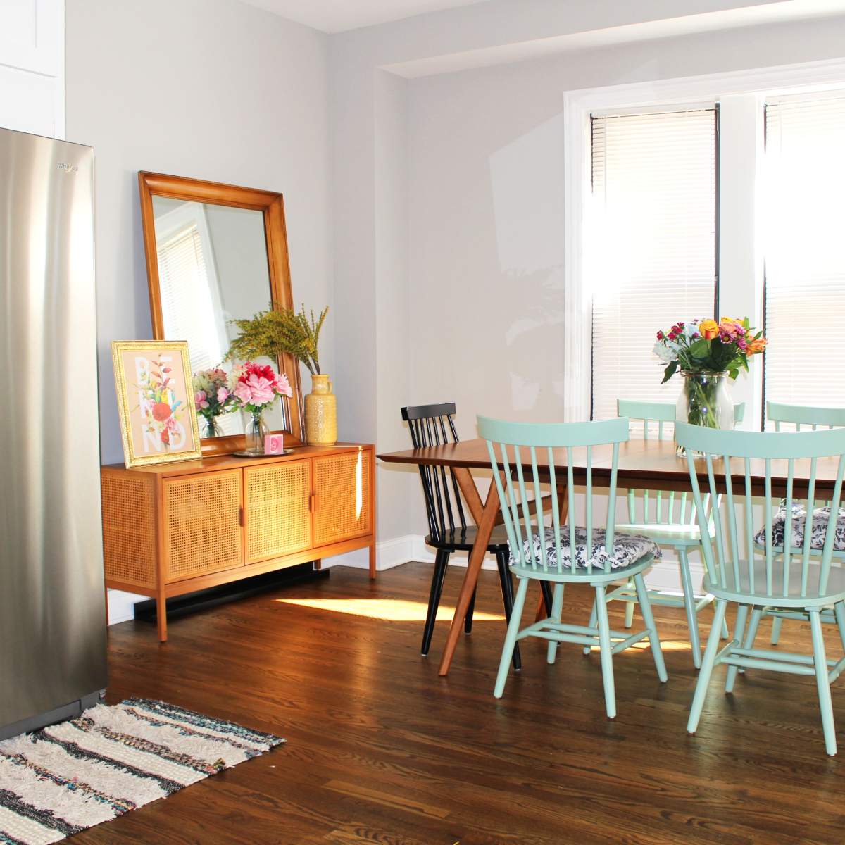 A modern kitchen scene features a stainless steel refrigerator, a wooden dining table surrounded by chairs, and a large mirror on the wall, with natural light coming through the windows.