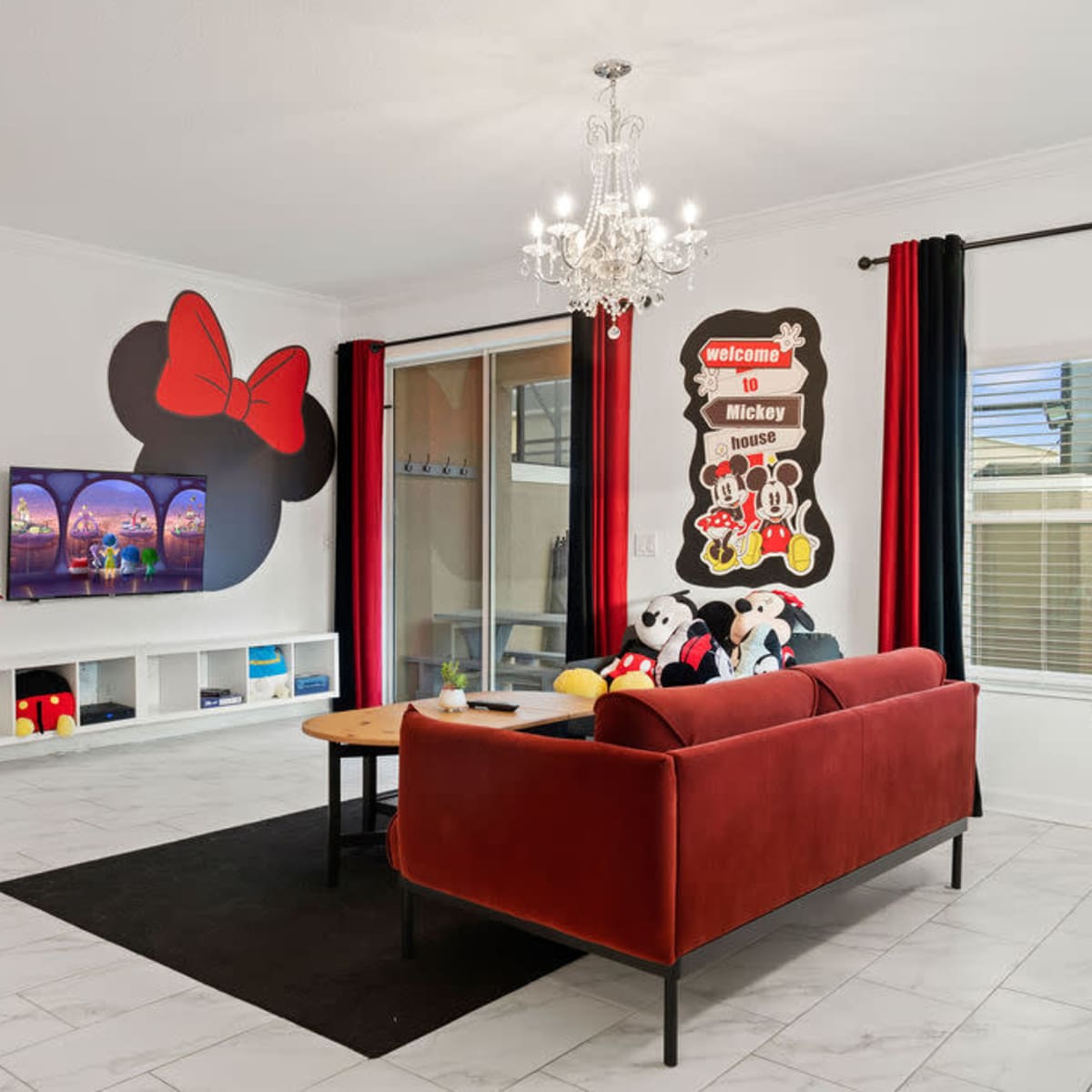 A living room decorated with Mickey Mouse-themed artwork, featuring a red couch, a large television, and playful accents like stuffed toys and colorful decor.