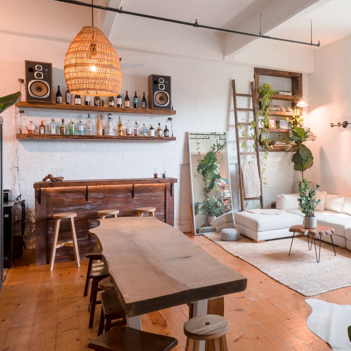 A bright, modern living area featuring a white sofa, a wooden dining table, a bar with bottles, and various indoor plants.