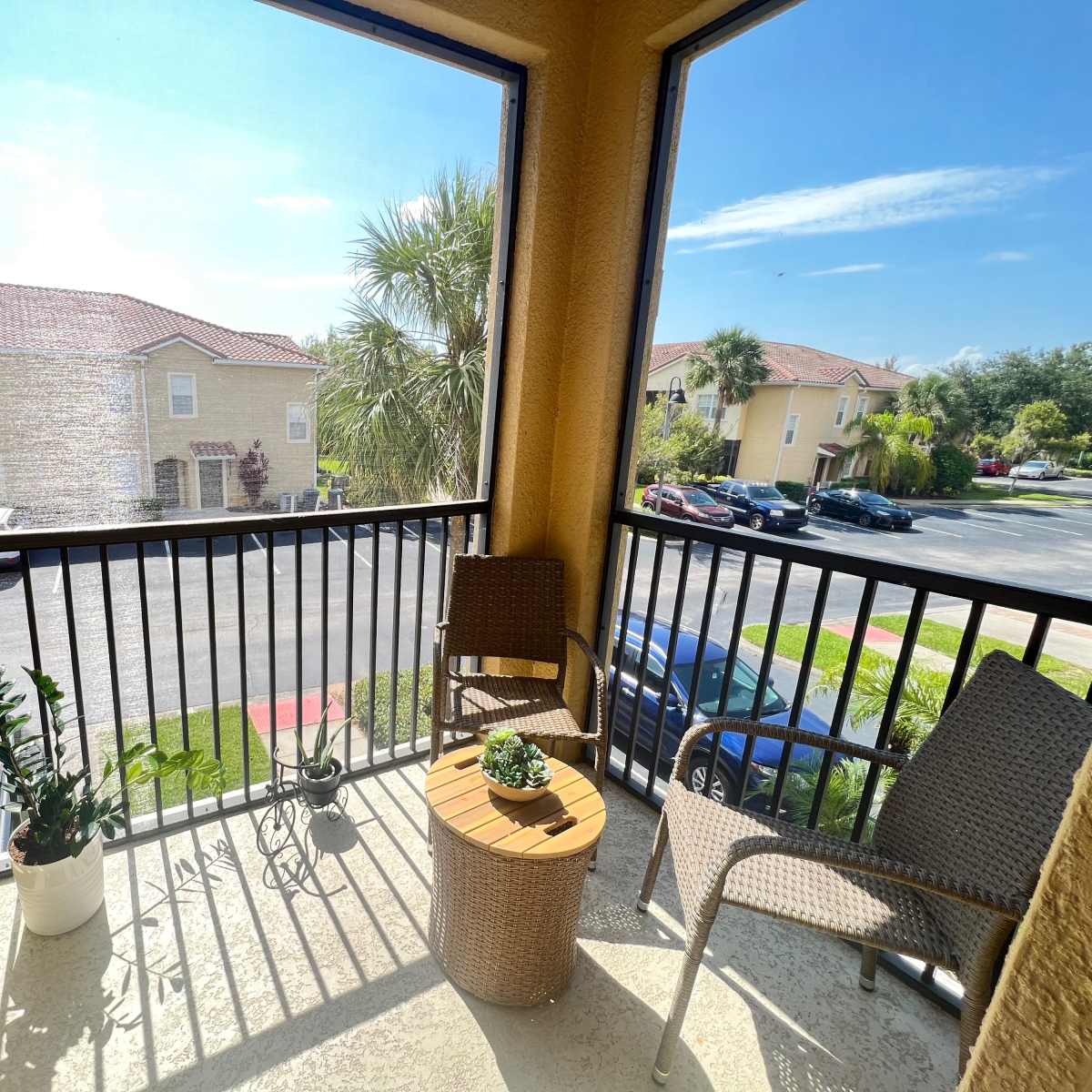 A small balcony with two chairs, a round table, and potted plants, overlooking a parking area and nearby buildings.