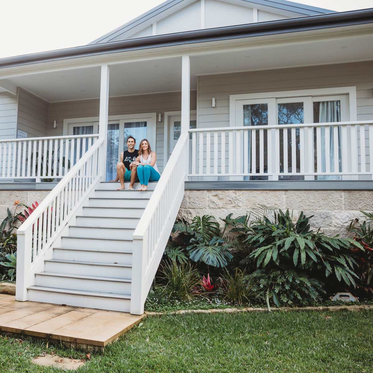 A couple sits on the steps of a large house with a well-maintained yard and vegetation.