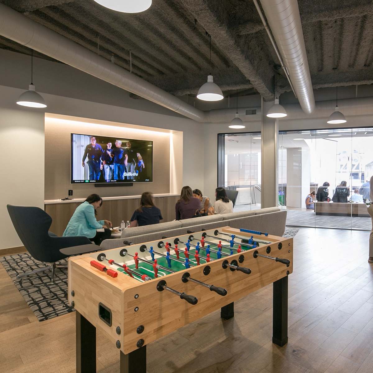 A common area features a foosball table in the foreground, with people engaged in various activities, such as watching a screen and socializing around tables.