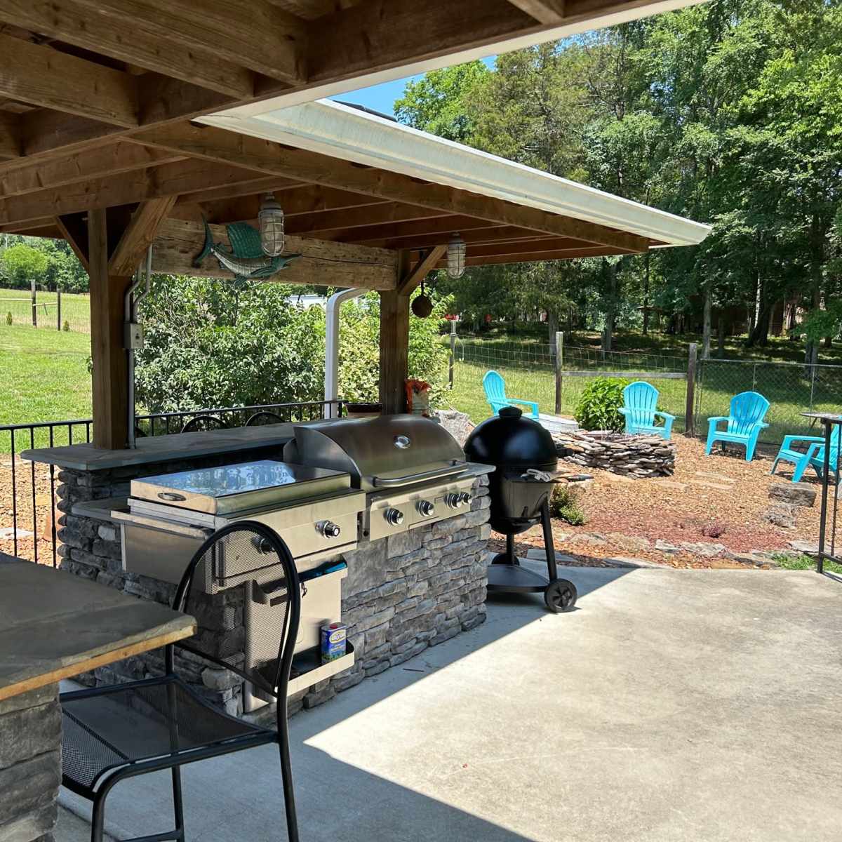An outdoor cooking area with a stone barbecue grill, seating area, and a view of a grassy lawn and seating chairs in the background.
