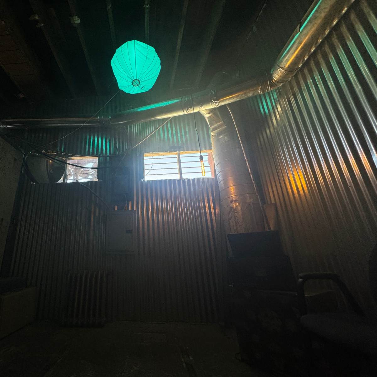 A dimly lit room with corrugated metal walls, a green paper lantern hanging from the ceiling, and a small window emitting light.