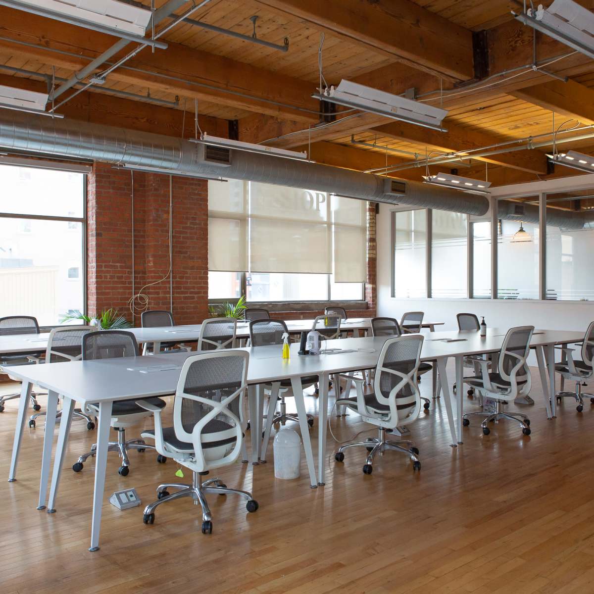 A modern office space with wooden floors, large windows, and several rows of desks with ergonomic chairs.