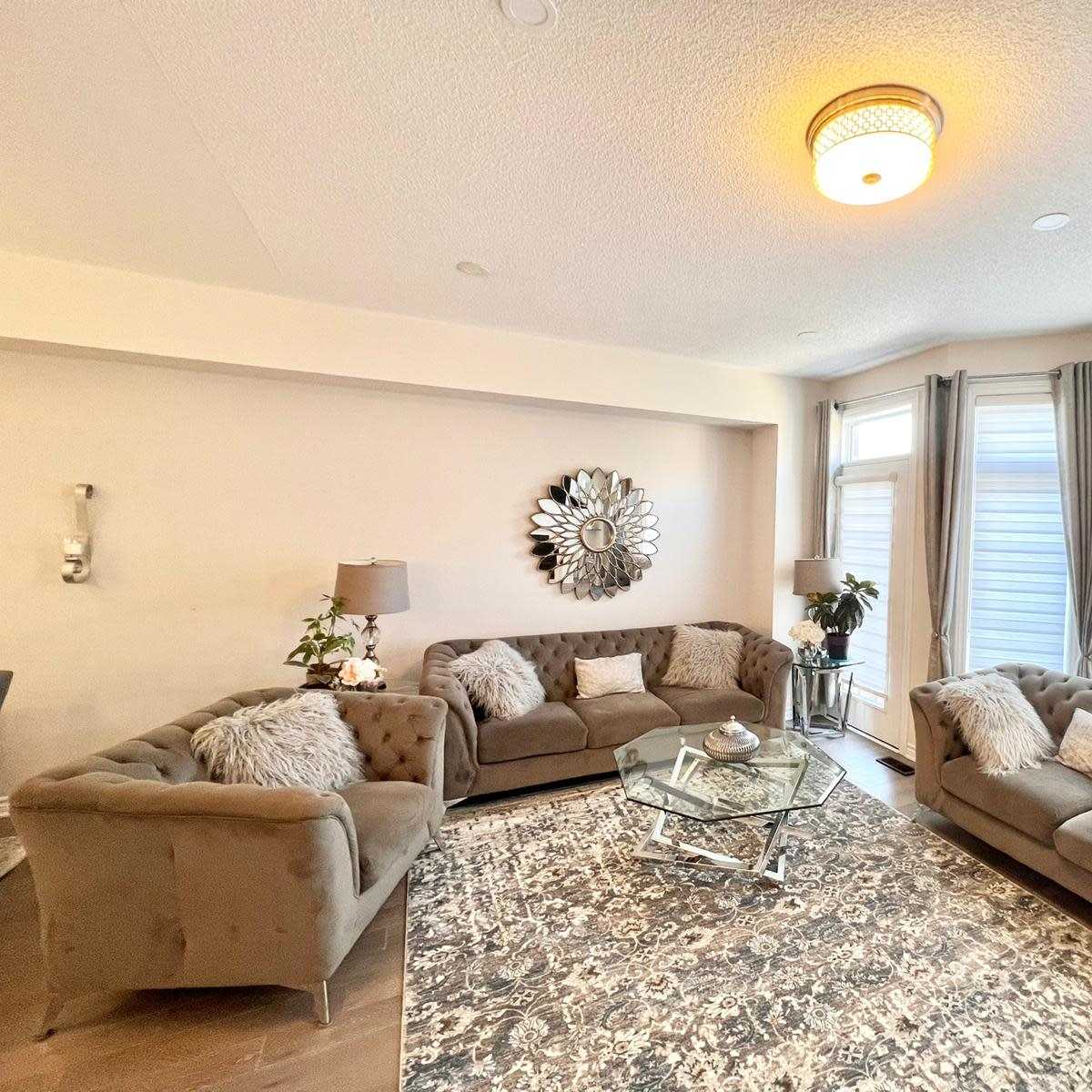 A modern living room features two plush sofas arranged around a central glass coffee table on a patterned rug, with large windows draped in curtains.