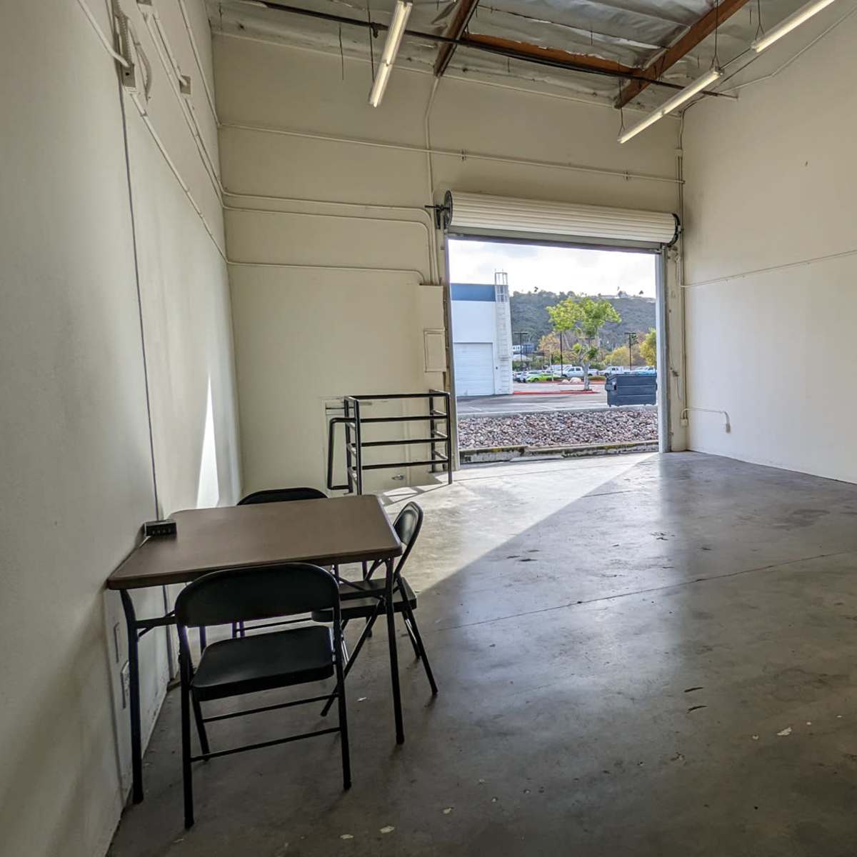 An empty industrial space with concrete flooring, a large open doorway, and a few pieces of furniture including a table and chairs.