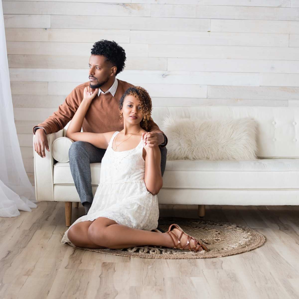 A couple sits together in a well-lit room, with the woman resting on the floor beside a sofa and the man comfortably positioned behind her.